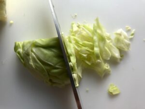 Knife slicing cabbage thin for Asian Coleslaw. It has cabbage, carrots, scallions, and sesame seeds in a creamy dressing with mayonnaise. vinegar, soy sauce, and Sesame Oil. It really is a sidekick that can go with anything!