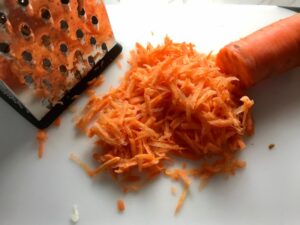 Shredding carrot on a grater for Asian Coleslaw. It has cabbage, carrots, scallions, and sesame seeds in a creamy dressing with mayonnaise. vinegar, soy sauce, and Sesame Oil. It really is a sidekick that can go with anything!