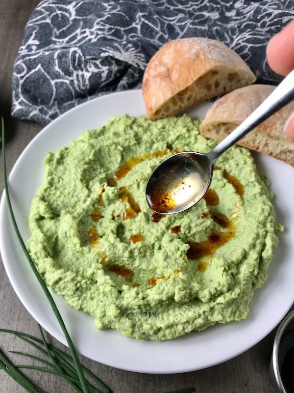 Spoon drizzling Harissa oil over Edamame Dip with Gorgonzola spread on a plate. It's creamy, savory, nutty, and utterly delicious!