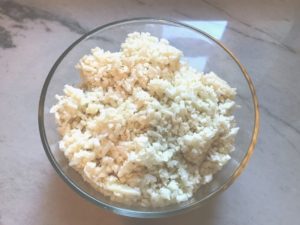 Raw Cauliflower Rice in a clear glass bowl on counter for Cheesy Cauliflower Rice recipe.