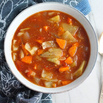 Detox Cabbage Soup in a bowl on blue napkin on counter. The soup is tomato-based with chunky veggies: cabbage, tomato, carrots, onion, and garlic.