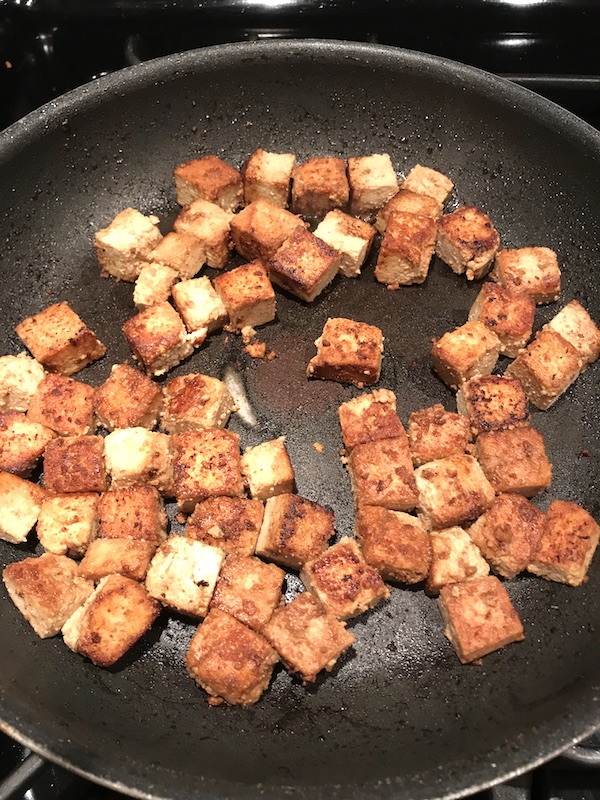 Cubes of marinated tofu being browned in a pan for Ginger Teriyaki Tofu bowl with noodles, zucchini strips, red peppers.