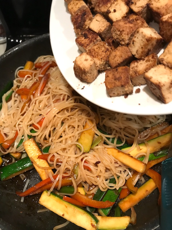 Browned tofu cubes being added back to pan with noodles and veggies for Ginger Teriyaki Tofu bowl with noodles, zucchini strips, red peppers.
