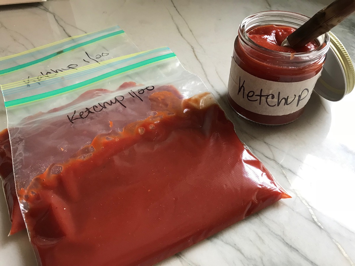 Jar labeled ketchup with spoon in it next to two storage baggies of ketchup also labeled for Best Honey Ketchup recipe.