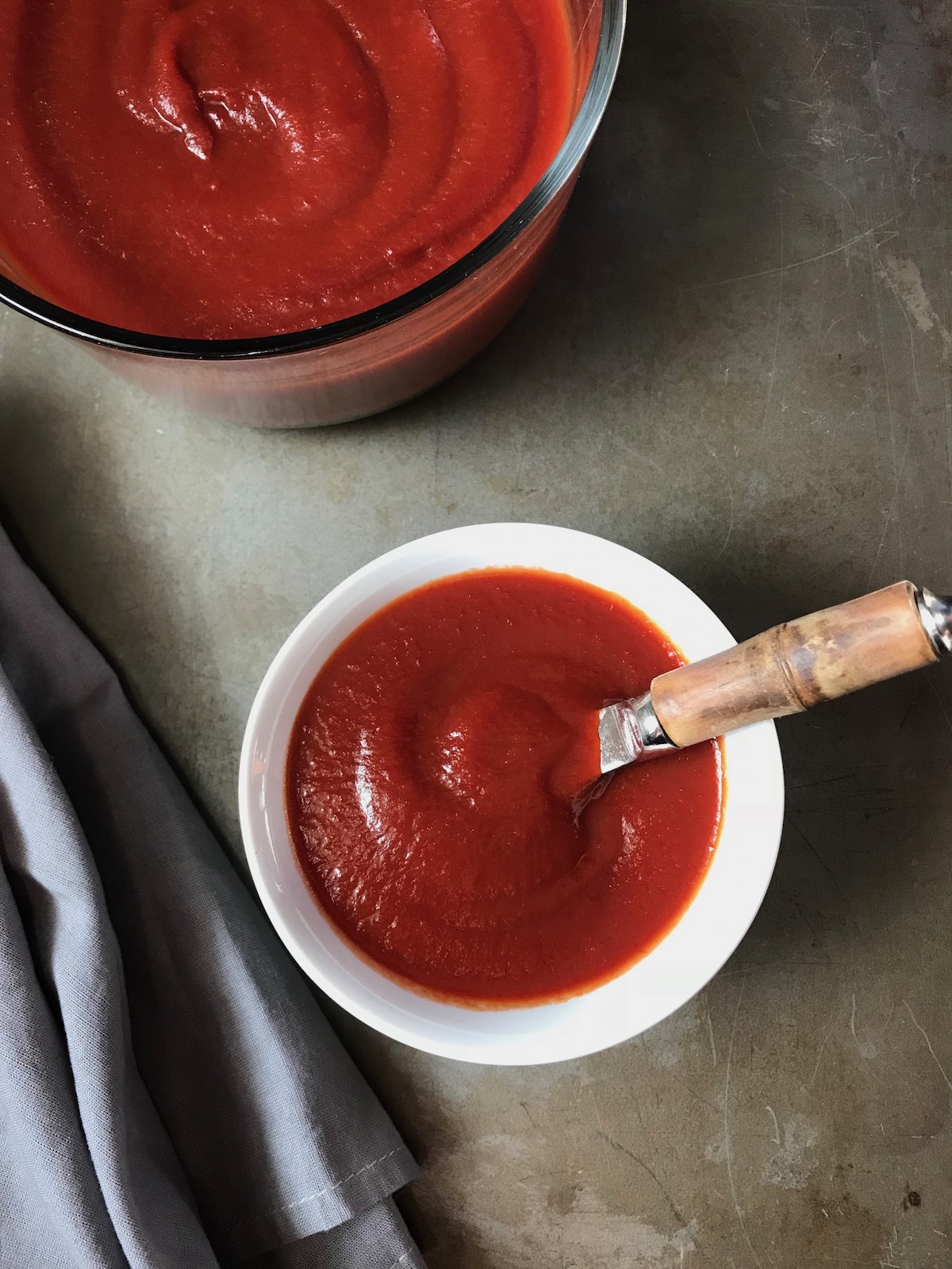 Homemade Honey Ketchup in a white serving bowl with spreading knife and large bowl of ketchup in background.