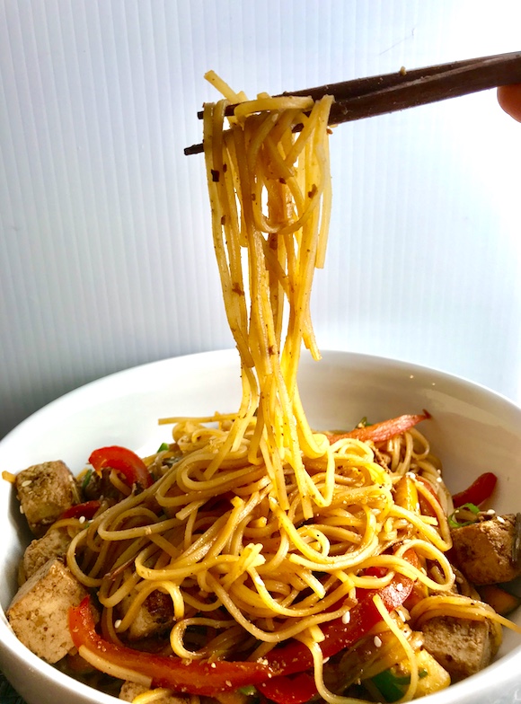 Chopsticks pulling up noodles from a bowl of Ginger Teriyaki Tofu with noodles, zucchini strips, red peppers, sesame seeds.