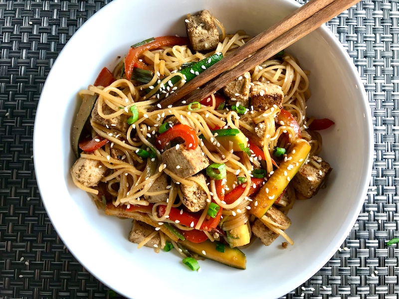 Ginger Teriyaki Tofu bowl with noodles, zucchini strips, red peppers, sesame seeds and chopsticks in the bowl.