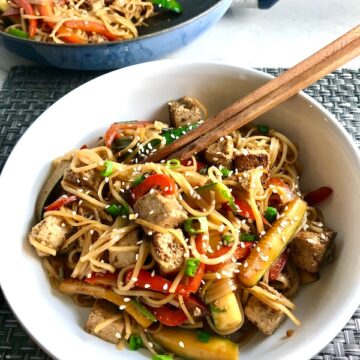 Ginger Teriyaki Tofu bowl with noodles, zucchini strips, red peppers, sesame seeds and chopsticks in the bowl. Pan with more stir fry in the background.