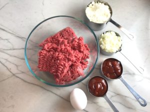 Raw Ground Beef in bowl with parmesan cheese, onions and ketchup in measuring cups on counter and egg on counter. This 5-Ingredient Easy Meatloaf recipe may be short on ingredients, but it is loaded with flavor. The lack of breadcrumbs means you don't have them soaking up the juices, so the meat stays moist - and it's gluten free too! Parmesan cheese, onion, egg, ketchup, salt, and pepper are all that go into this yummy loaf of goodness!