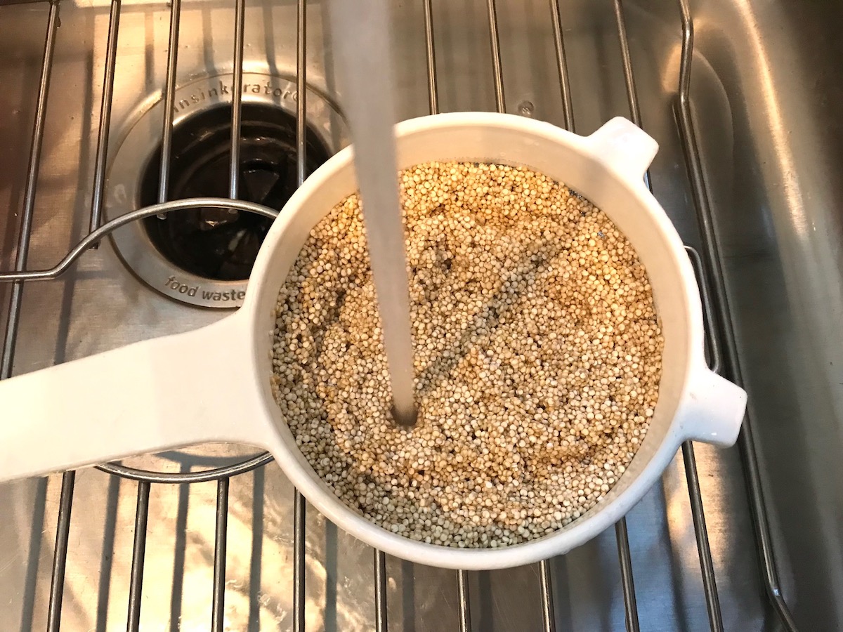 Uncooked white quinoa in a strainer with running water rinsing it in over sink for Creamy Parmesan Mushroom Quinoa Risotto