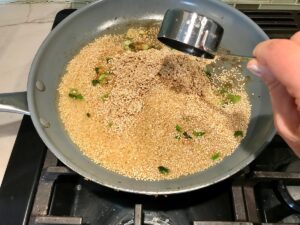Hand adding 1 measuring cup of broth to quinoa in frying pan with scallions and garlic for Creamy Parmesan Mushroom Quinoa Risotto.