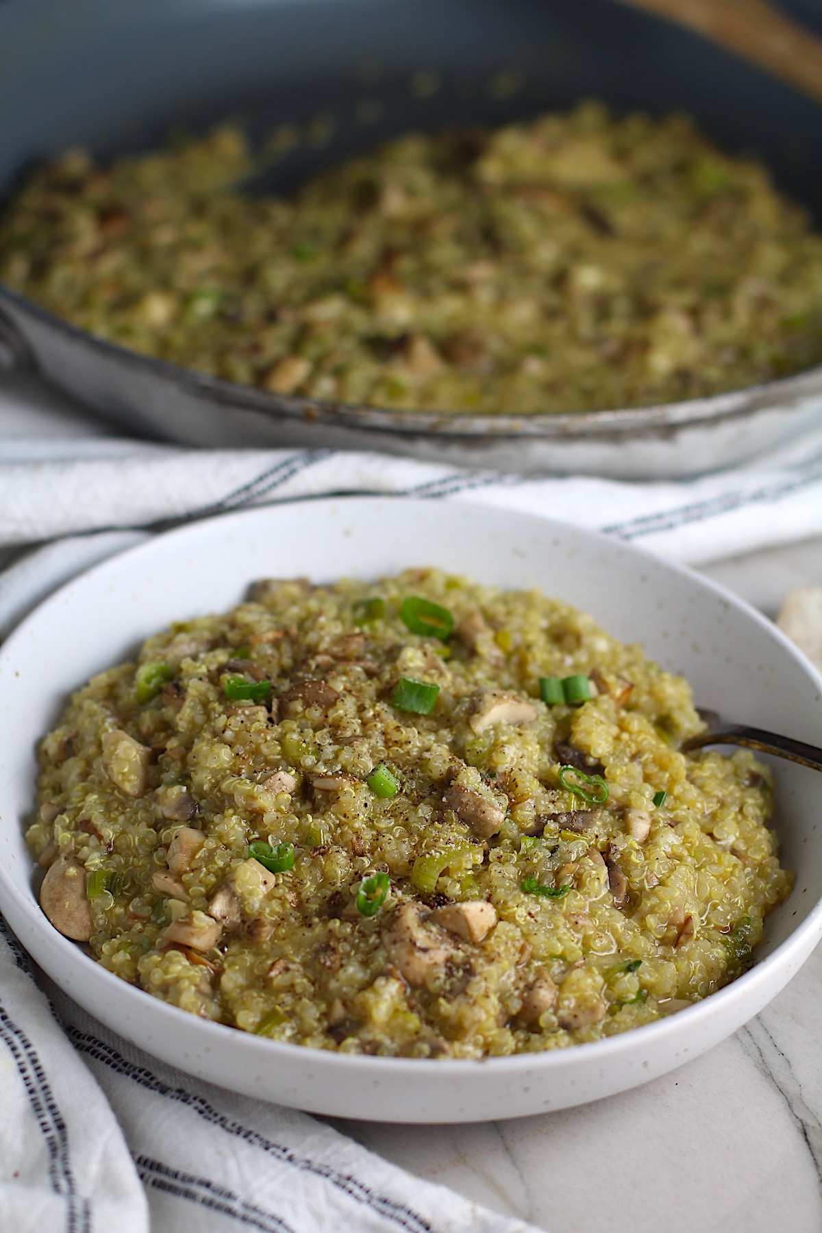 Creamy Parmesan Mushroom Quinoa Risotto in a bowl with fork and skillet in background. This recipe has the traditional garlic flavor in the background and the nuttiness from the Parmesan combined with the meaty earthy flavor of the mushrooms. There is no wine.