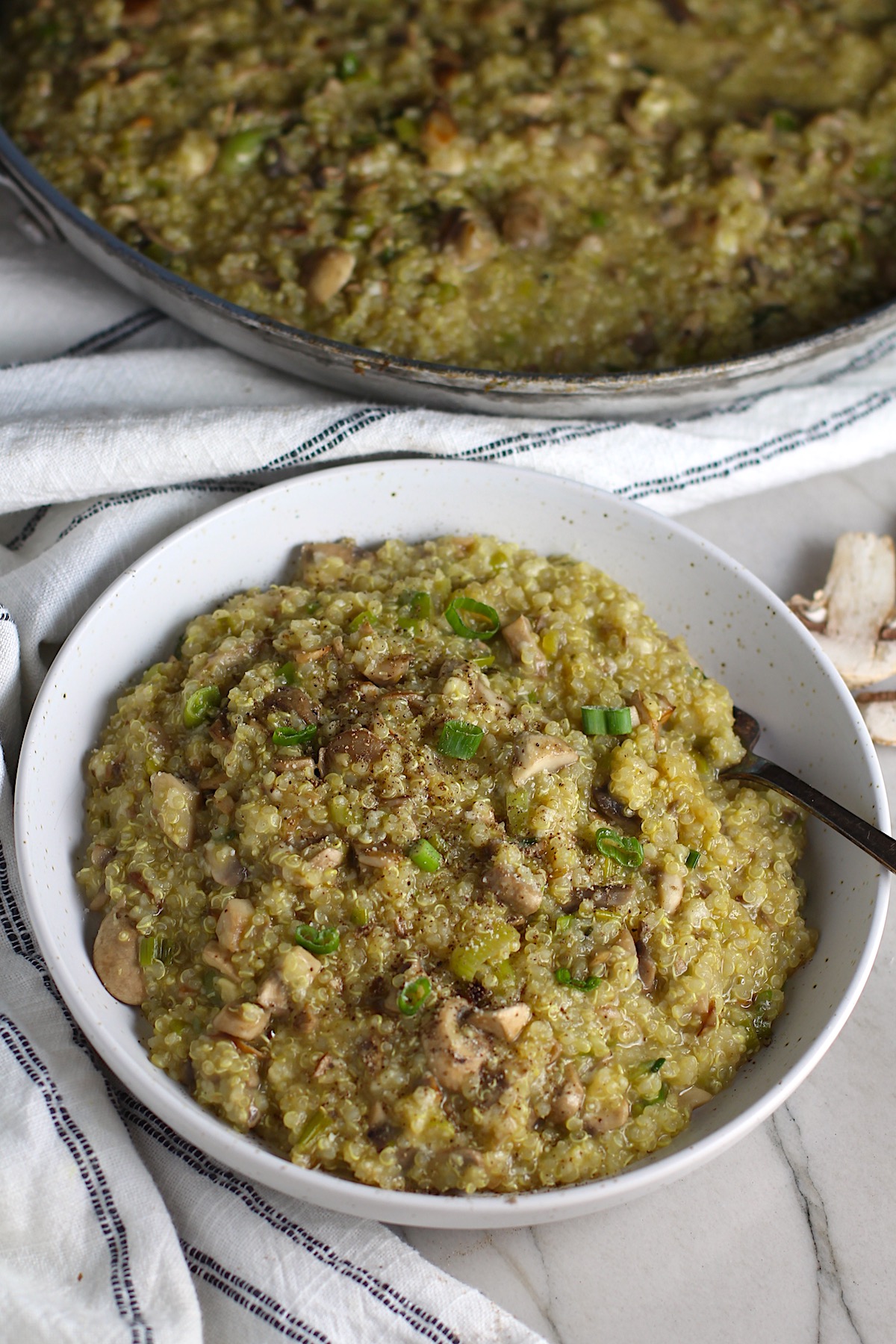 Creamy Parmesan Mushroom Quinoa Risotto in a bowl with fork and skillet in background. This recipe has the traditional garlic flavor in the background and the nuttiness from the Parmesan combined with the meaty earthy flavor of the mushrooms. There is no wine.