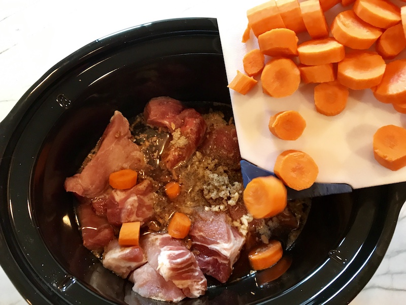 Sliced uncooked carrots on a cutting board being poured into a slow cooker with raw pork and other ingredients.