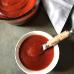Best Sugar Free Ketchup in a white serving bowl with spreading knife and large bowl of ketchup in background.