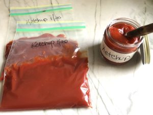 Whisk mixing honey and tomato paste in pot for the Best Sugar Free Ketchup in a storage bag with jar of ketchup to the right..