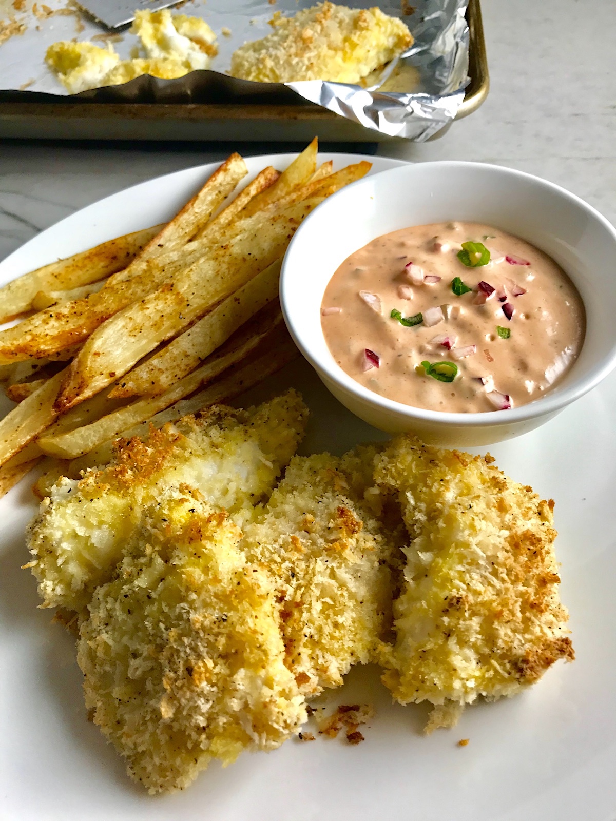 Crispy Baked Cod Panko Fish and Chips on a plate with side of remoulade dipping sauce and pan in background. Everything bakes in the oven on a sheet pan to save time and energy.