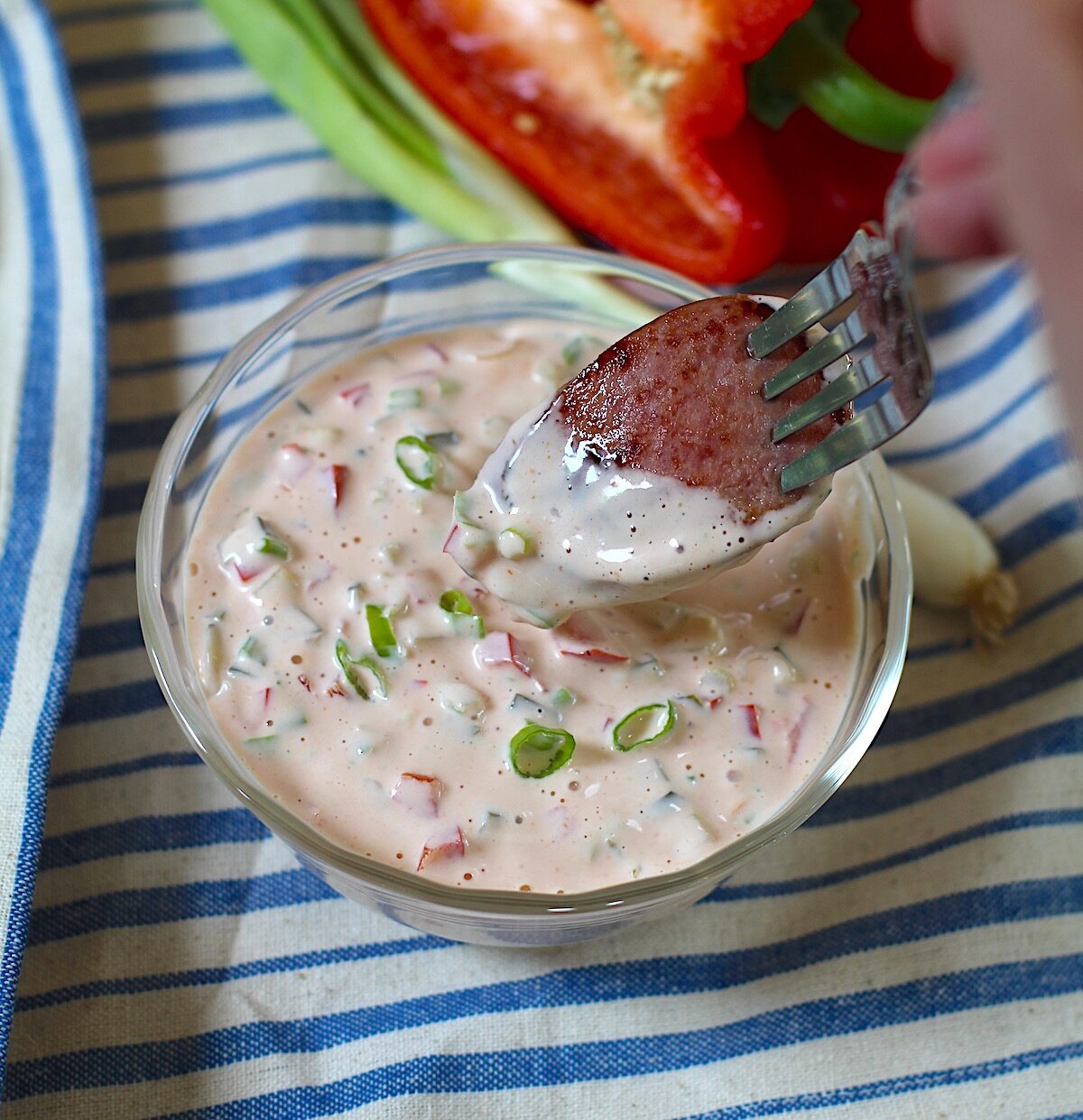 Fork dipping a slice of sausage into Crunchy Mayo and Ketchup dipping sauce with pickles, red peppers, and scallions on a blue and white striped towel with whole scallion and cut red pepper sitting next to it..