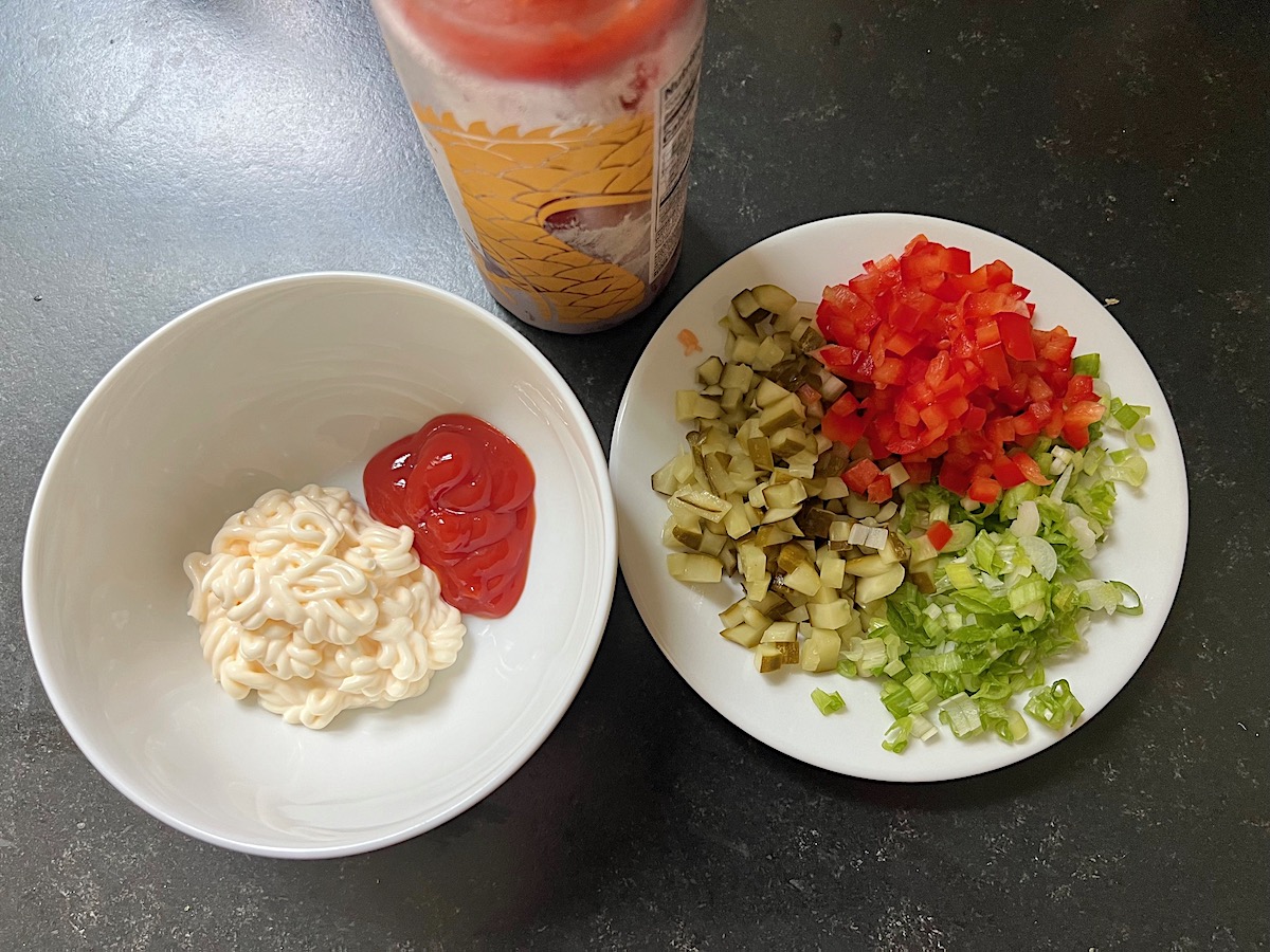 Ingredients in bowls for Mayo and Ketchup Remoulade Sauce: left bowl has mayo (left) ketchup (right). Right bowl has chopped pickles, red peppers, scallions. In back on counter is a bottle of Sriracha sauce.