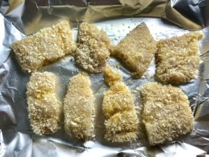 Raw cod fish breaded on a sheet pan lined with aluminum before being baked for Crispy Baked Cod Panko Fish and Chips.
