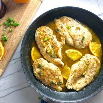 Orange Brown Butter Sauce Chicken with orange slices and parsley garnish on top in skillet on counter with wood cutting board in background with sliced oranges and parsley.