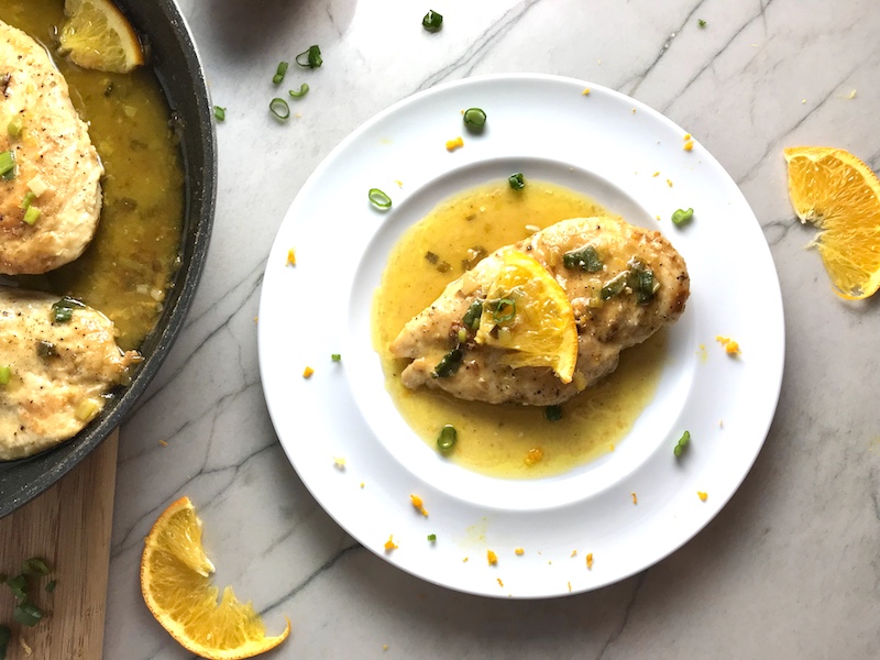 One piece of Orange Brown Butter Sauce chicken on a white plate with an orange slice and parsley garnish on top. The plate is on the counter and next to it is the pan of chicken with orange slices also on the counter.