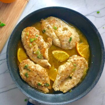 Orange Butter Sauce Chicken with orange slices and parsley garnish on top in skillet on counter with wood cutting board in background with sliced oranges and parsley.