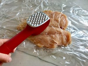 Hand using meat tenderizer to pound out chicken breast.