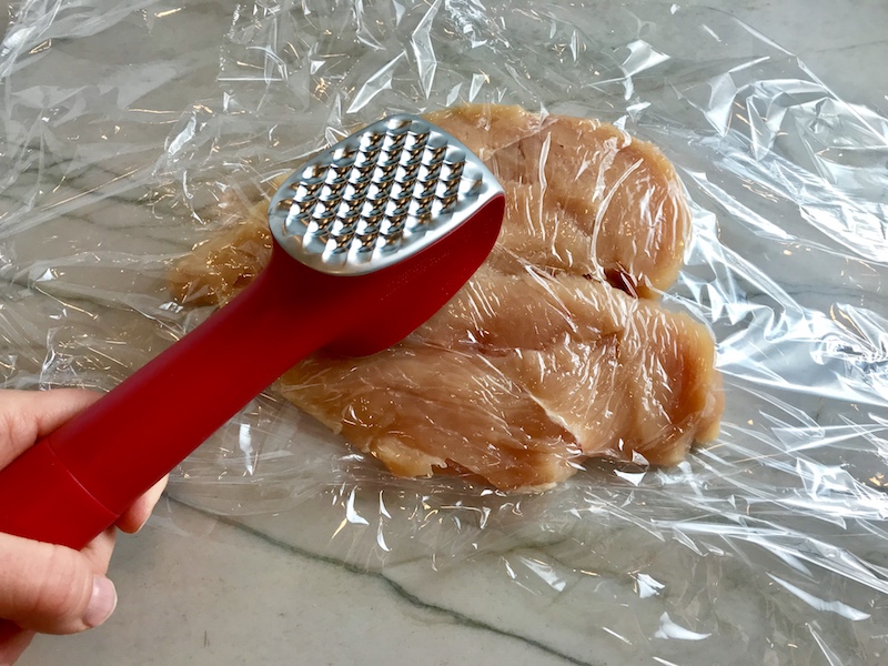 Hand using meat tenderizer to pound out chicken breast.
