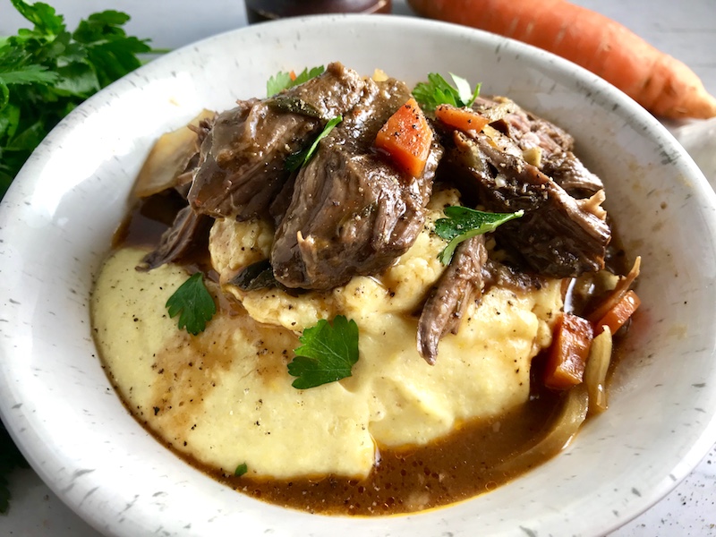 Easy Slow Cooker Short Ribs Cooked short ribs on top of creamy polenta in a bowl with sauce, carrots, and onions for flanken short ribs recipe slow cooker. Bowl sitting on counter.