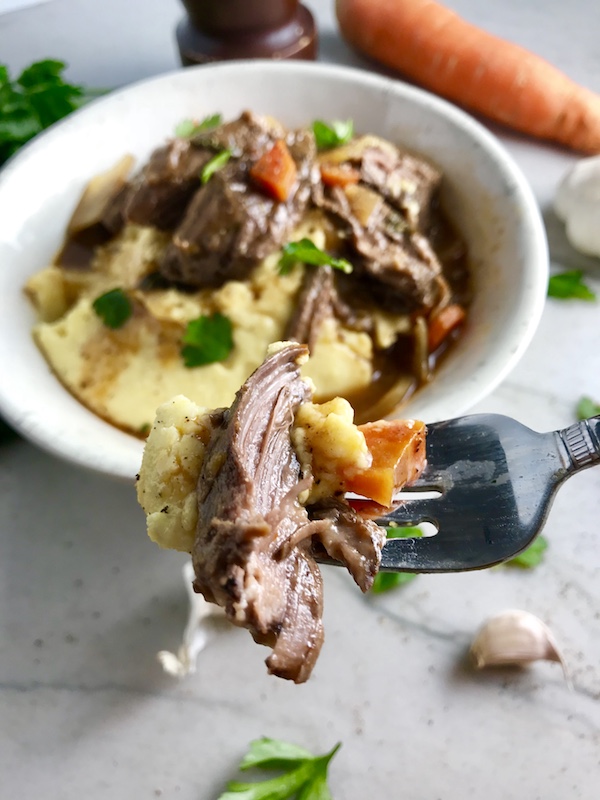 Easy Slow Cooker Short Ribs - fork tender Cooked short ribs on top of creamy polenta in a bowl with sauce, carrots, and onions for flanken short ribs recipe slow cooker. Bowl sitting on counter.