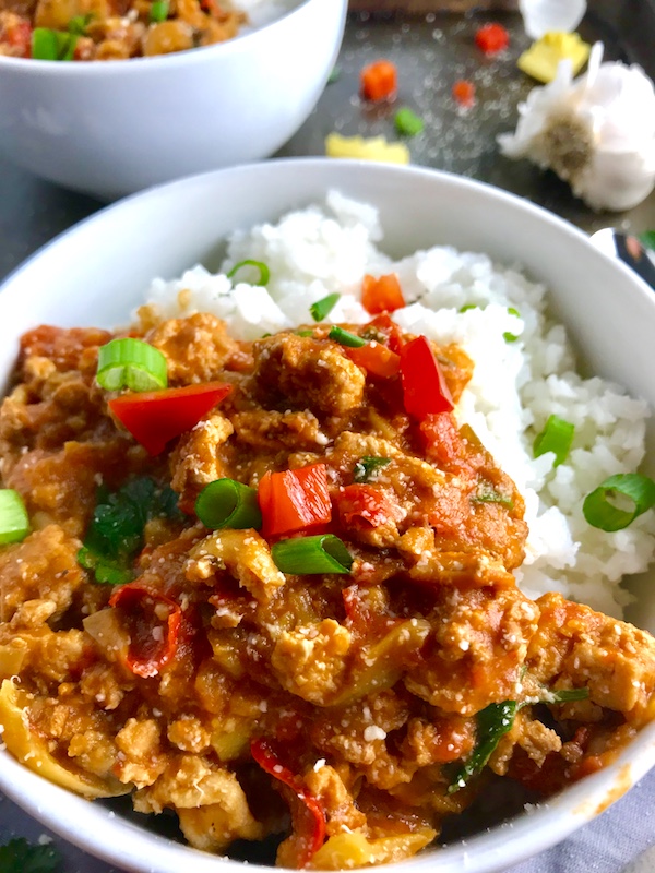 Slow cooker Ground chicken ragu in a bowl over rice with diced red pepper, scallions, and pieces of artichoke and garlic cloves on counter with a spoon.