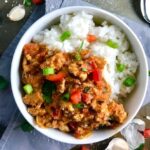 Slow cooker Ground chicken ragu in a bowl over rice with diced red pepper, scallions, and pieces of artichoke and garlic cloves on counter with a spoon.