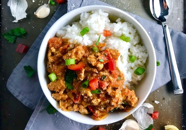 Slow cooker Ground chicken ragu in a bowl over rice with diced red pepper, scallions, and pieces of artichoke and garlic cloves on counter with a spoon.