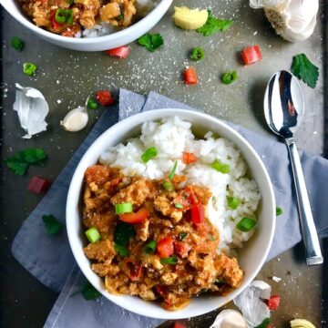 Slow cooker Ground chicken ragu in a bowl over rice with diced red pepper, scallions, and pieces of artichoke and garlic cloves on counter with a spoon.