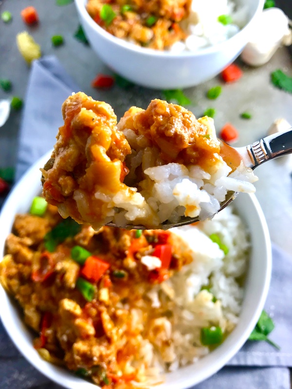 Slow cooker Ground chicken ragu in a bowl over rice with diced red pepper, scallions, and pieces of artichoke and garlic cloves on counter with a spoon.