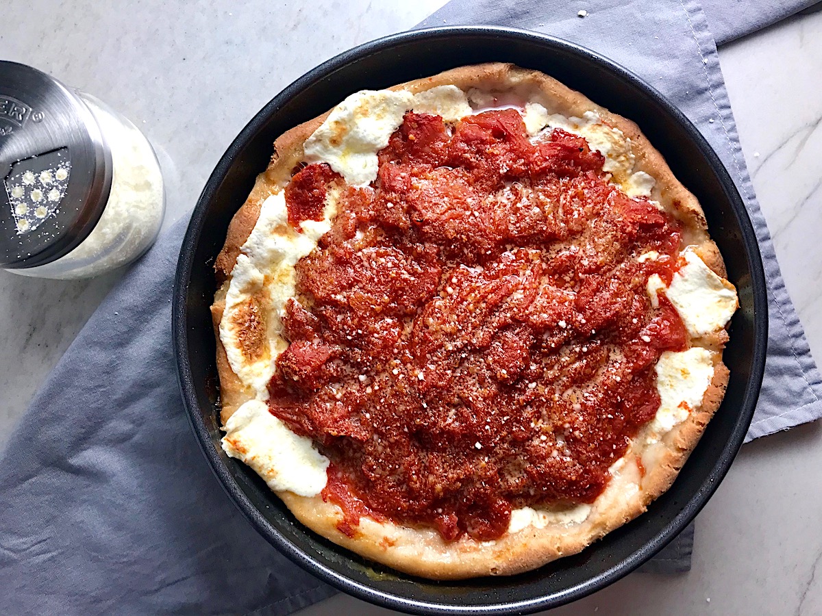 Chicago Style deep dish pizza with chicken sausage and tomatoes on top in a round pan on a towel on counter with grated parmesan cheese next to it..