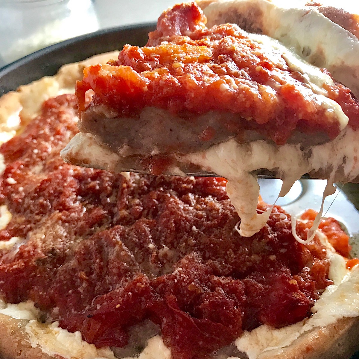 Spatula lifting slice Chicago Style deep dish pizza with chicken sausage and tomatoes on top from a round pan.