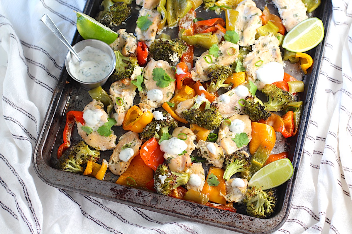 Cilantro Lime Yogurt Sauce for Chicken with Peppers and Broccoli cooked on a sheet pan with sauce drizzled over top and lime wedges on the side.