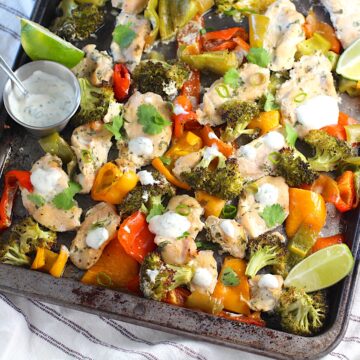 Cilantro Lime Yogurt Sauce for Chicken with Peppers and Broccoli cooked on a sheet pan with sauce drizzled over top and lime wedges on the side.