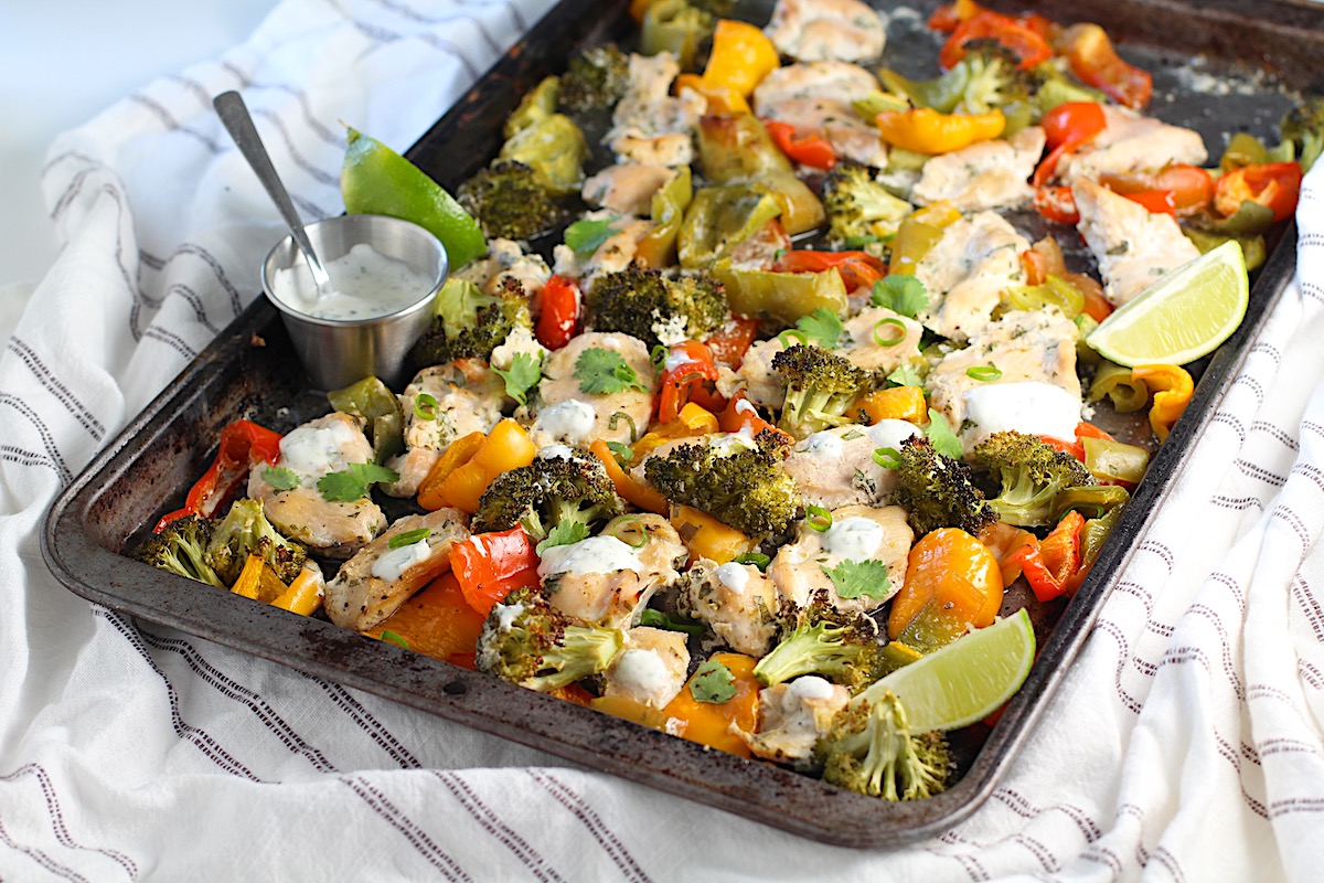 Cilantro Lime Yogurt Sauce for Chicken with Peppers and Broccoli cooked on a sheet pan with sauce drizzled over top and lime wedges on the side.