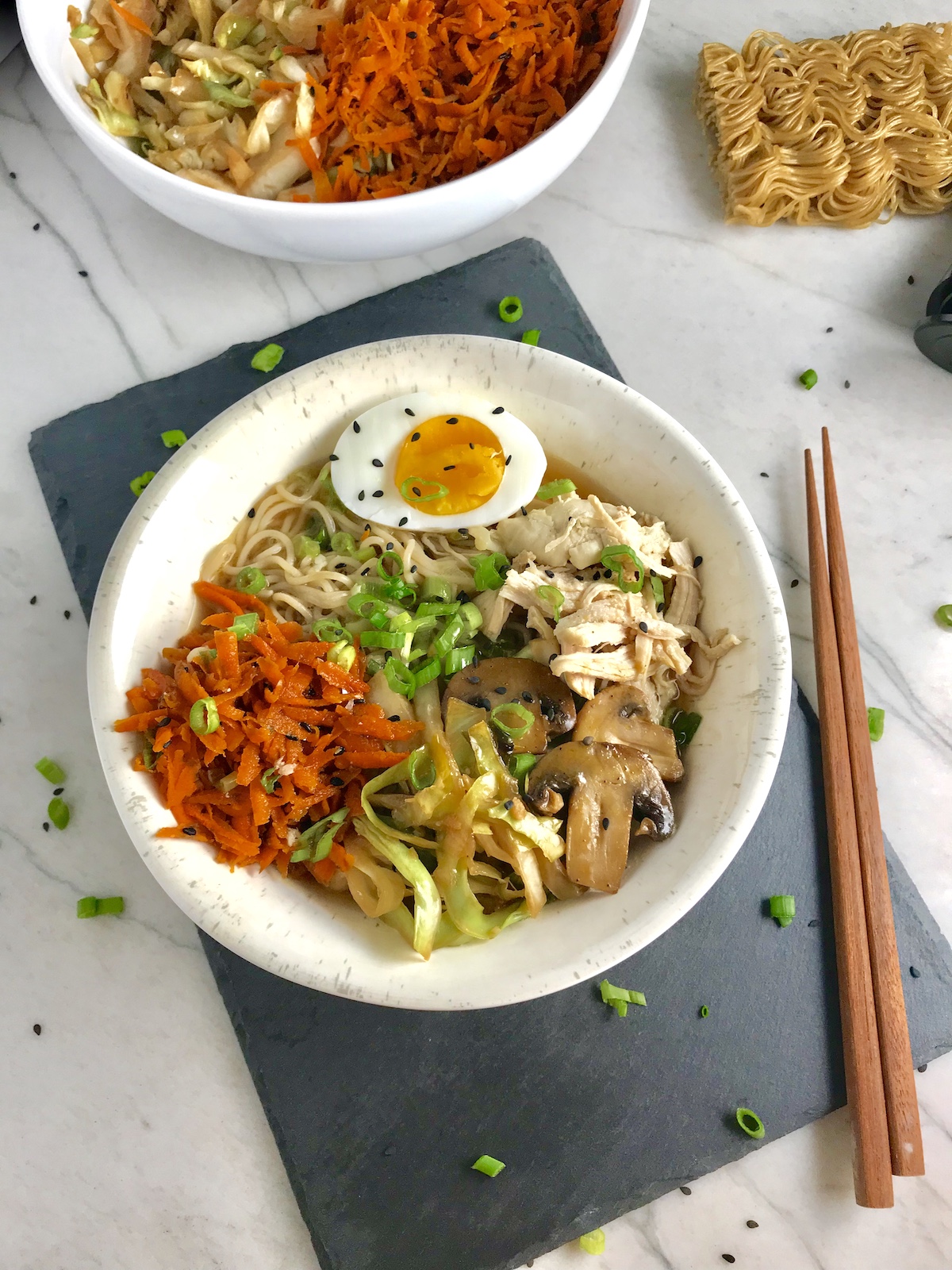 Asian Noodle Bowl with Chicken, carrots, cabbage, half of a hard boiled egg, scallions, and black sesame seeds with chopsticks on the side.
