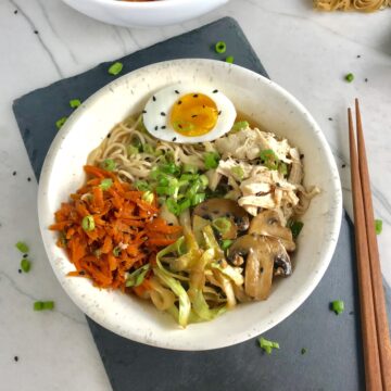 Healthy Chicken Ramen Noodles with Chicken, carrots, cabbage, half of a hard boiled egg, scallions, and black sesame seeds with chopsticks on the side.