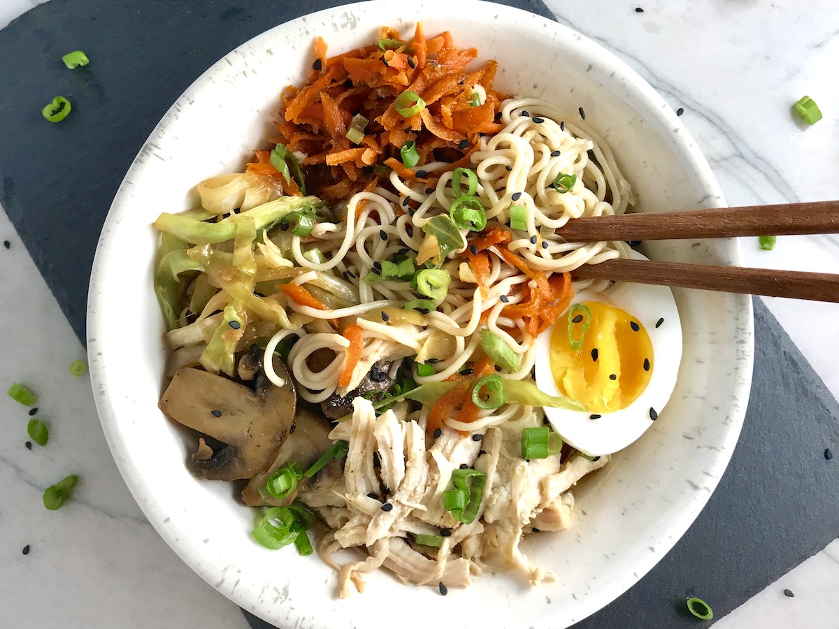Asian Noodle Bowl recipe in a bowl with chopsticks. The bowl has Chicken, carrots, cabbage, half of a hard boiled egg, scallions, and black sesame seeds.