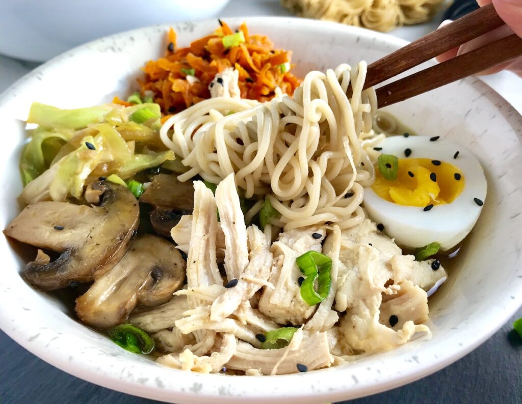 Healthy Chicken Ramen Noodles in a bowl with chopsticks. The bowl has Chicken, carrots, cabbage, half of a hard boiled egg, scallions, and black sesame seeds.