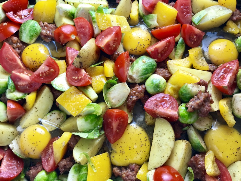 Veggies and raw eggs on pan for One Pan Italian Sausage and Eggs. It's LOADED with flavors and textures. You get the salty Italian Sausage, sweet bell pepper, creamy potatoes, sweet and juicy tomatoes, meaty brussel sprouts, creamy eggs, and salty, nutty cheddar cheese. Best of all, the entire meal cooks on one large pan!