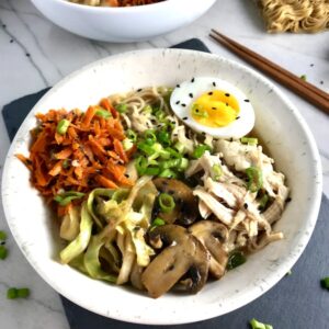 Healthy Chicken Ramen Noodles with Chicken, carrots, cabbage, half of a hard boiled egg, scallions, and black sesame seeds with chopsticks on the side.