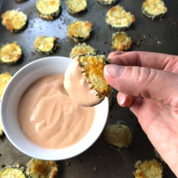 Breaded Zucchini Chips on pan with Sriracha dipping sauce in a bowl on pan. Hand dipping one zucchini chip into sauce.