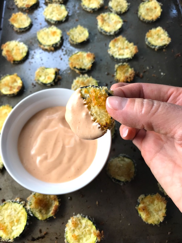 Breaded Zucchini Chips on pan with Sriracha dipping sauce in a bowl on pan. Hand dipping one zucchini chip into sauce.