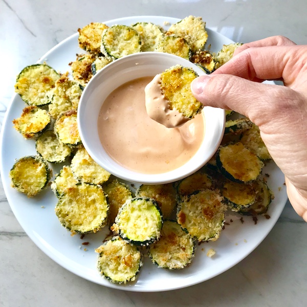 hand holding zucchini chip after dipping in sauce. Plate with chips and dip. Learn how to roast zucchini slices.